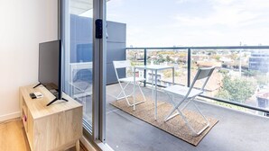 Un balcón con vistas a un paisaje urbano, un televisor sobre un soporte de madera y dos sillas blancas.