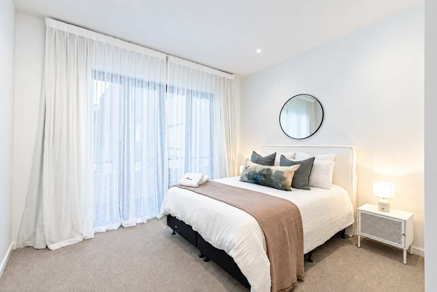 Quarto de hotel com cama, mesa de cabeceira, abajur, espelho e cortinas leves.