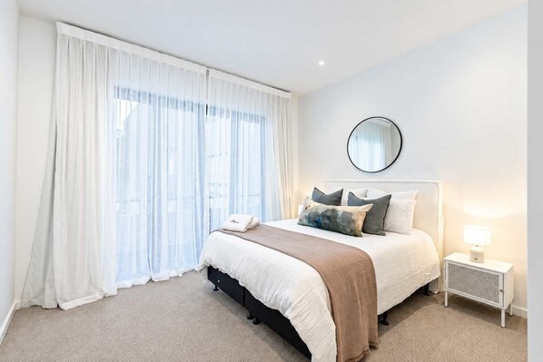 Une chambre d’hôtel avec un lit, une table de chevet, une lampe, un miroir et des rideaux légers.