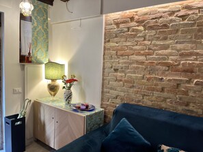 Ein Zimmer mit einem blauen Sofa, einem kleinen Tisch mit Lampe und Vase, einer Backsteinwand und einer Tür.