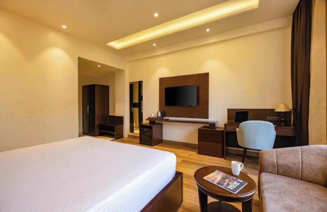 Una habitación de hotel con una cama grande, un escritorio con computadora, un televisor montado en la pared y una zona de estar con una silla y una mesa pequeña.