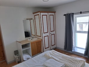 A bedroom with a bed, a wooden wardrobe, a mirror, a sink, and a window with curtains.