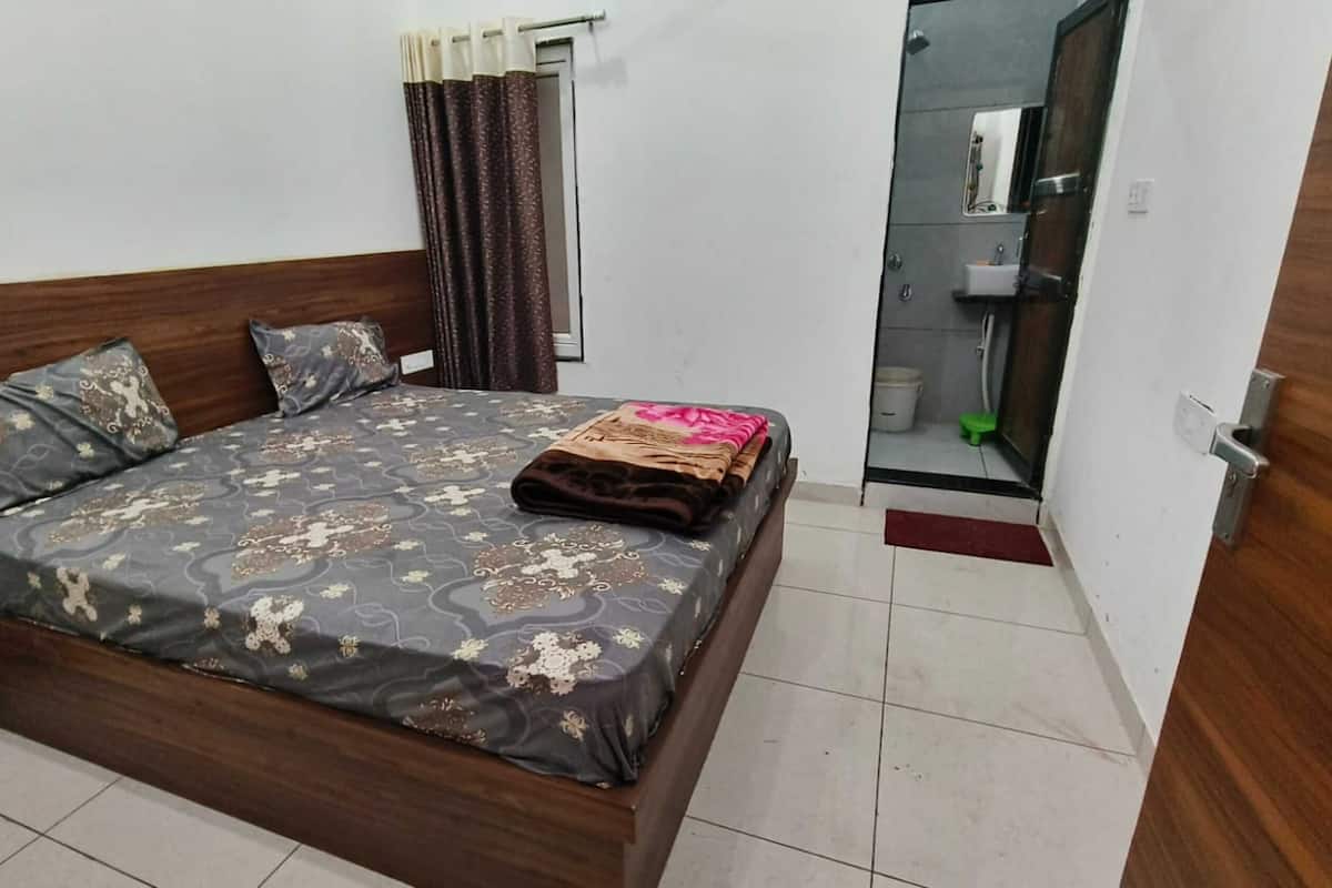 Un dormitorio con cama, almohadas, una colcha, ventana con cortinas, puerta que da a un baño y suelo de baldosas.
