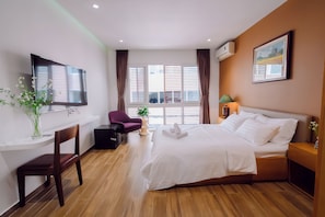 A hotel room with a bed, a TV, a desk, a chair, a window with blinds, a lamp, and a painting on the wall.