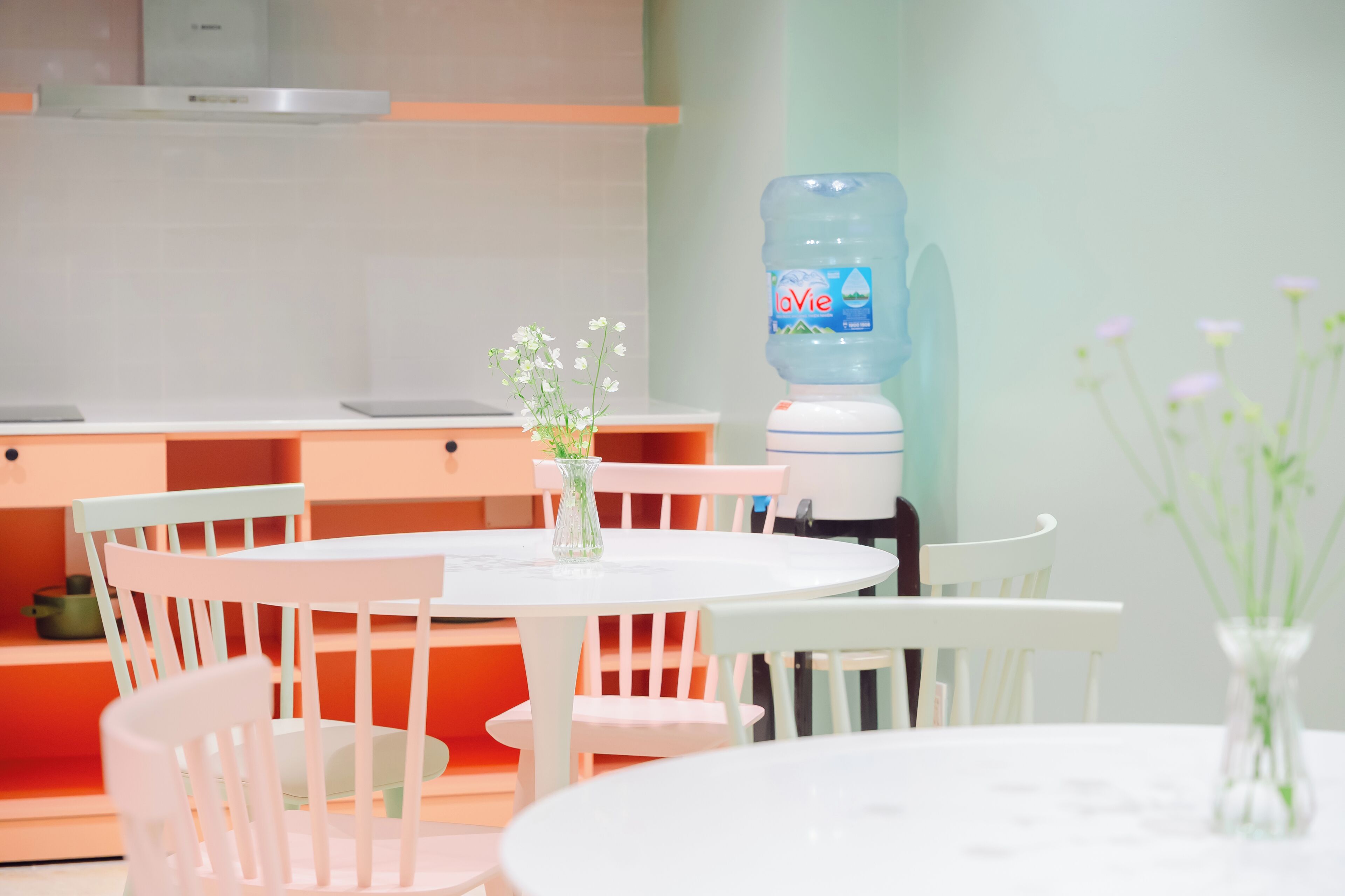 A dining area with white round tables and chairs, a water dispenser, and a vase with flowers.