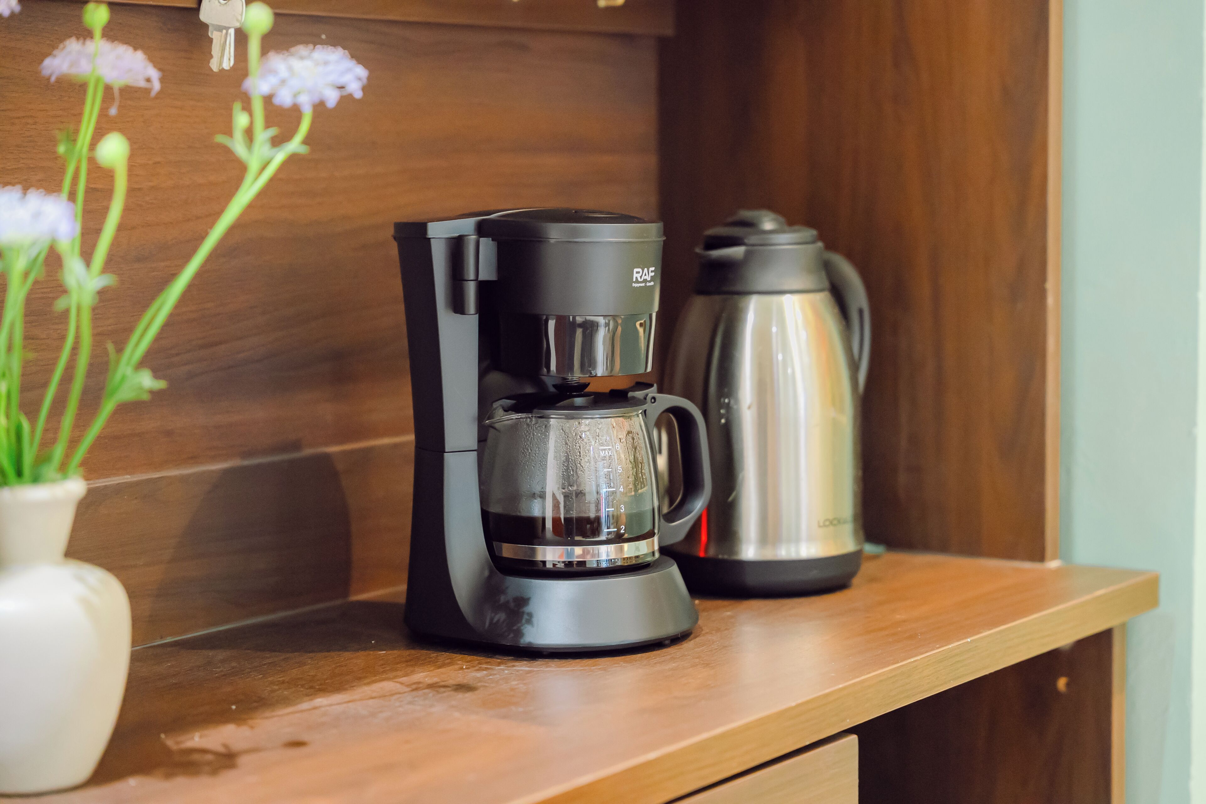 Una cafetera sobre una encimera de madera, un recipiente de agua de acero inoxidable y un jarrón blanco con plantas verdes.