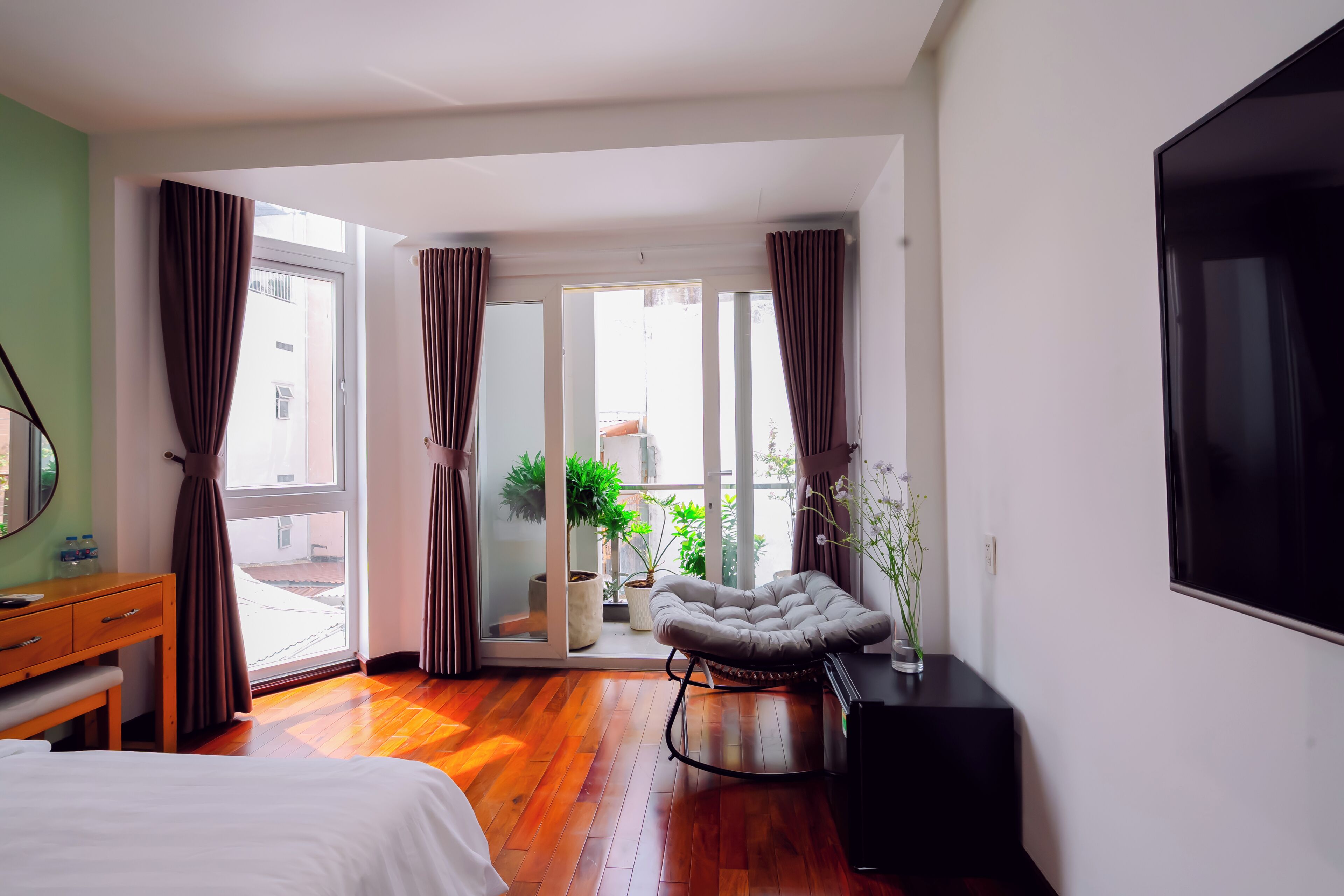 A hotel room with a bed, a desk, a chair, a TV, and a balcony with plants.