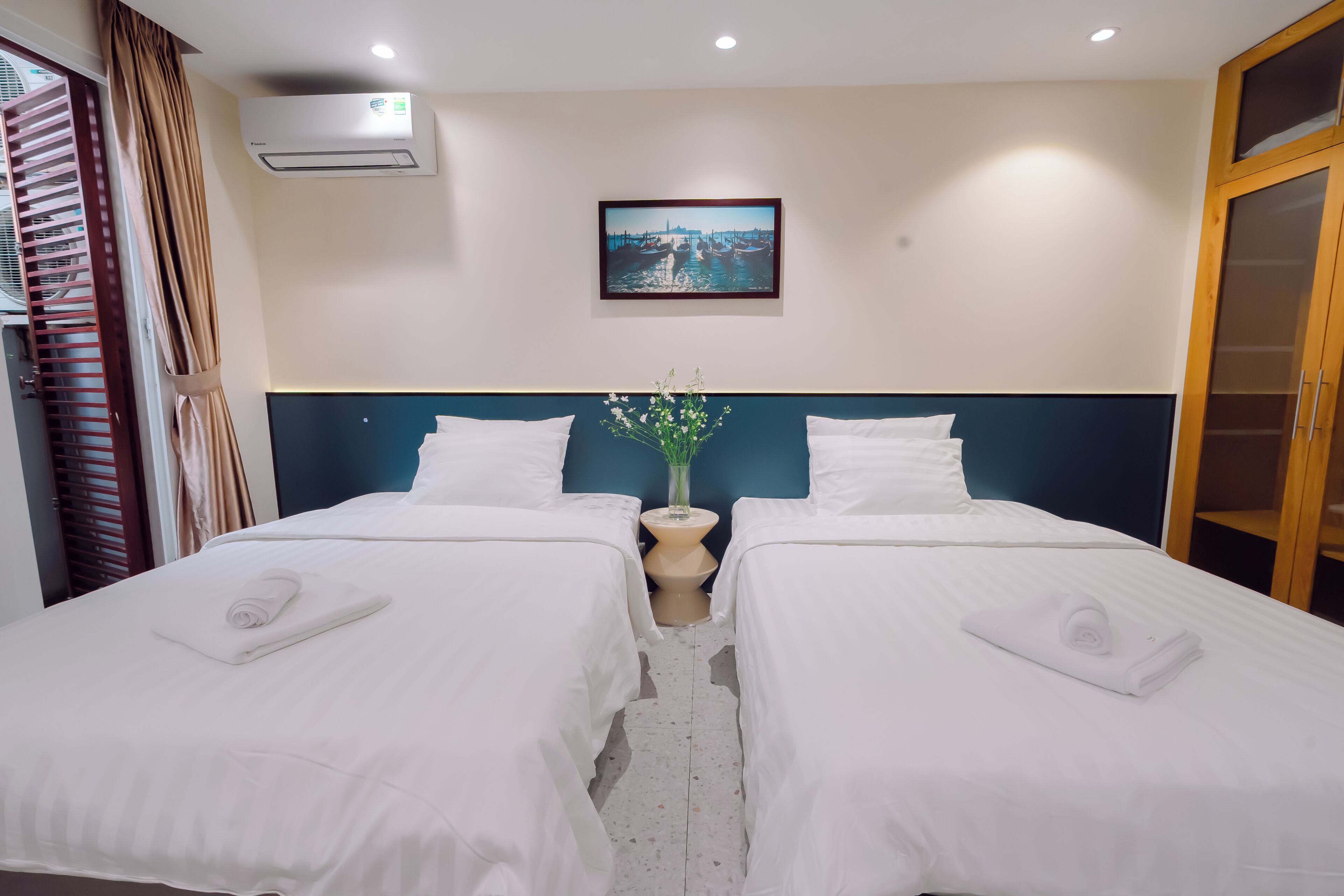 Quarto de hotel com duas camas, ar-condicionado fixado na parede, um quadro emoldurado e uma pequena mesa com um vaso.