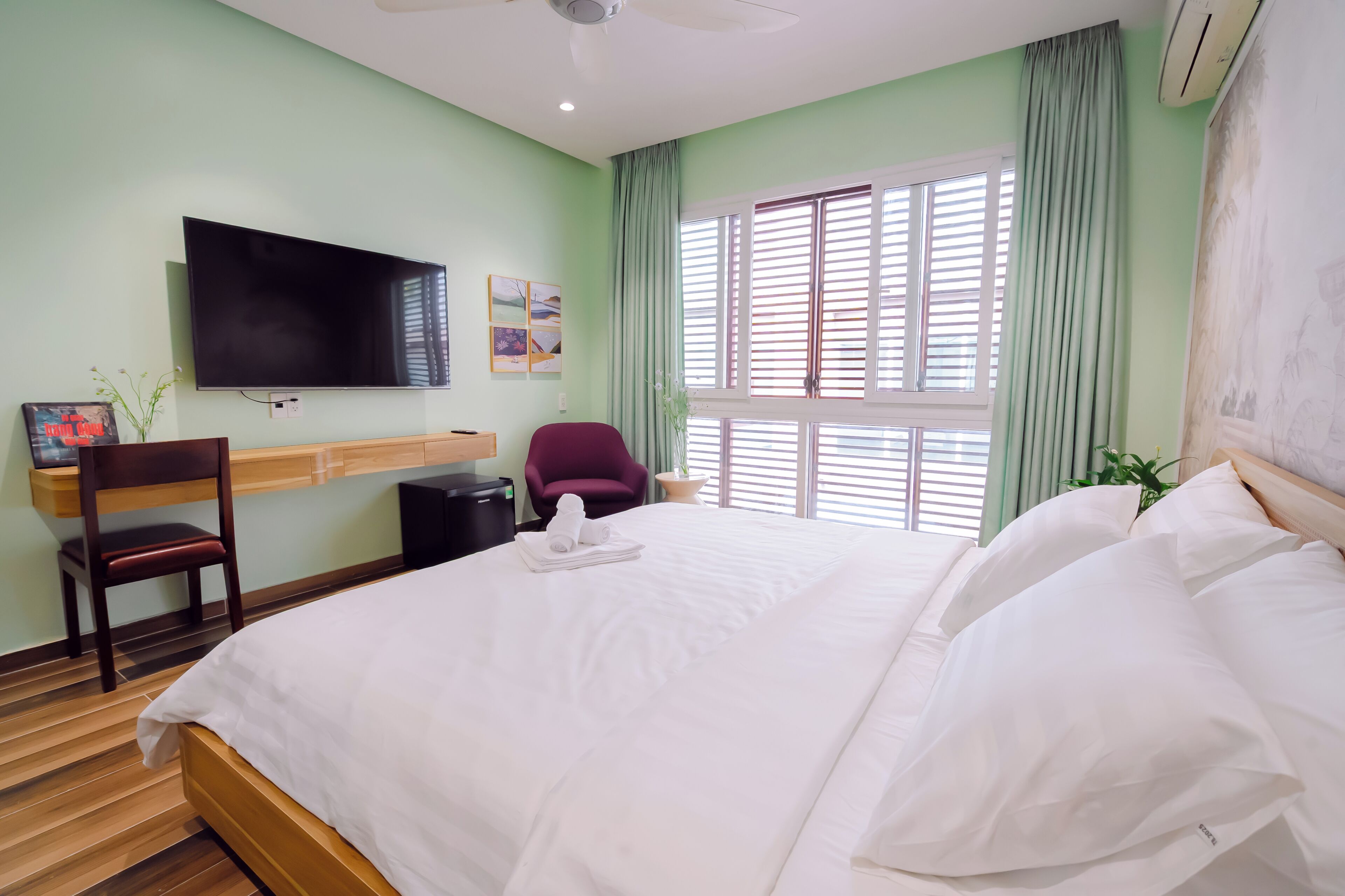 A hotel room with a bed, a desk with a chair, a TV mounted on the wall, and a window with blinds.