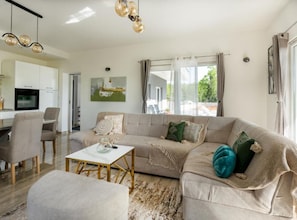 Een moderne woonkamer met een bank, salontafel en voetenbankje, een eethoek met stoelen, en een keuken met een televisie.