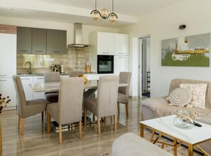 A modern kitchen with white cabinets, a dining area with a glass table, and a living room with a sofa and a painting on the wall.
