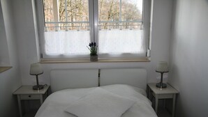 Un dormitorio con cama, dos mesitas de noche, una ventana con vistas y una planta pequeña en el alféizar.
