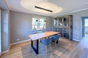 Een eethoek met een houten tafel, blauwe stoelen, hanglampen en een glazen kast met planken.