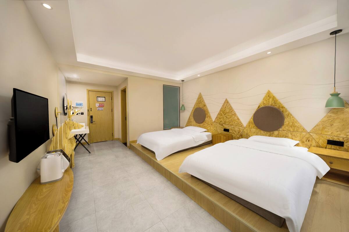 A hotel room with two beds, a TV, a wooden bench, a small table, and a wall-mounted lamp.