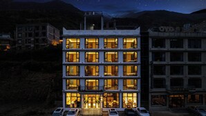 A multi-story hotel with illuminated windows and balconies, a parking lot with several cars, and a mountainous backdrop. - Jianshanxi Designer Hotel (Ngawa Autonomous Prefecture)