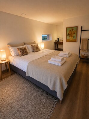 A neatly made bed with a beige blanket and pillows, a bedside table with a lamp, a small window, and a painting on the wall.