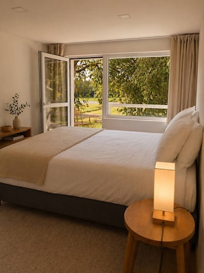 A bedroom with a large bed, a bedside table with a lamp, and a view of greenery through a window.