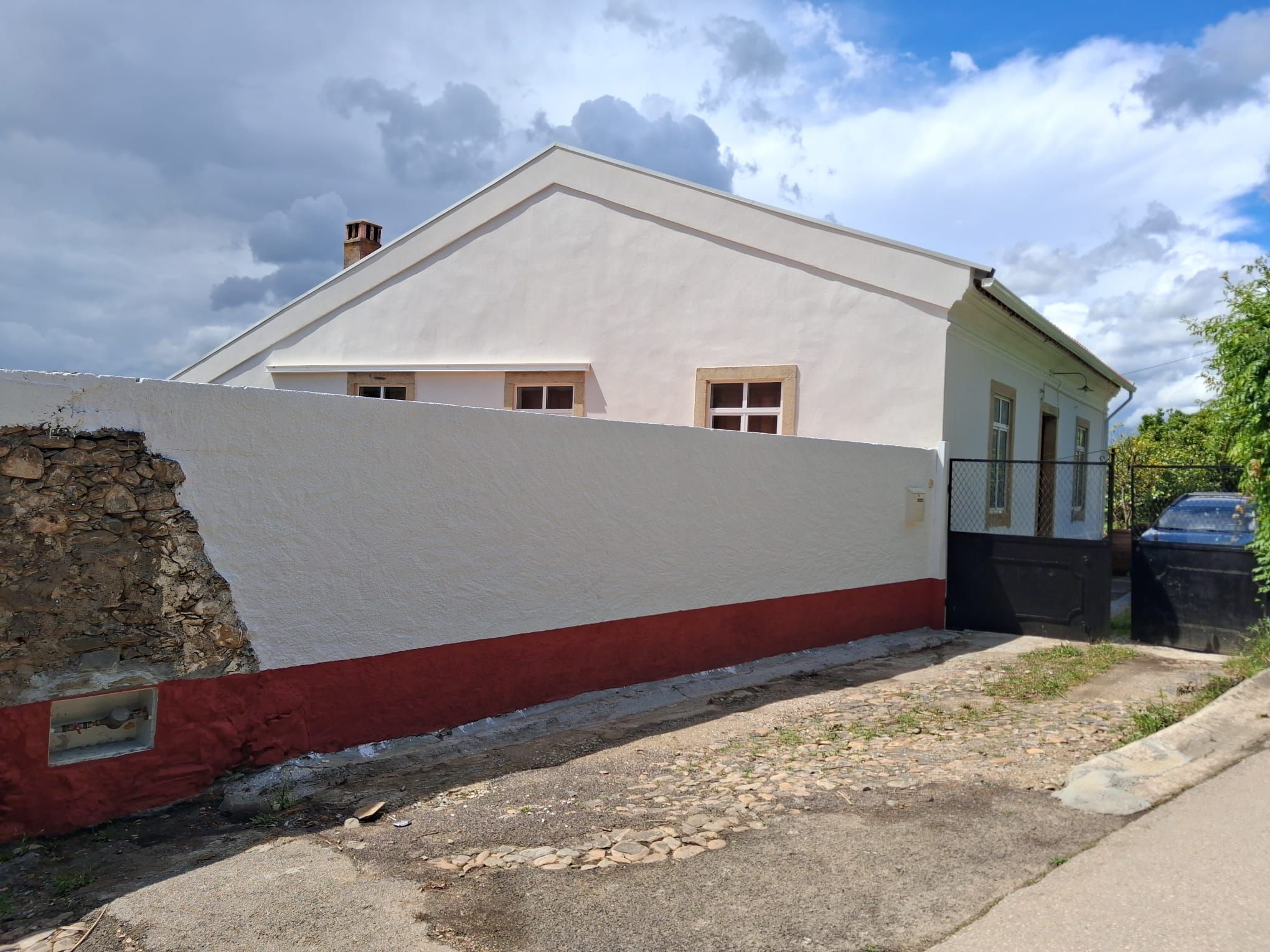 A single-story building with a white facade and a red base, a stone wall to the left, and a parked car in the background.
