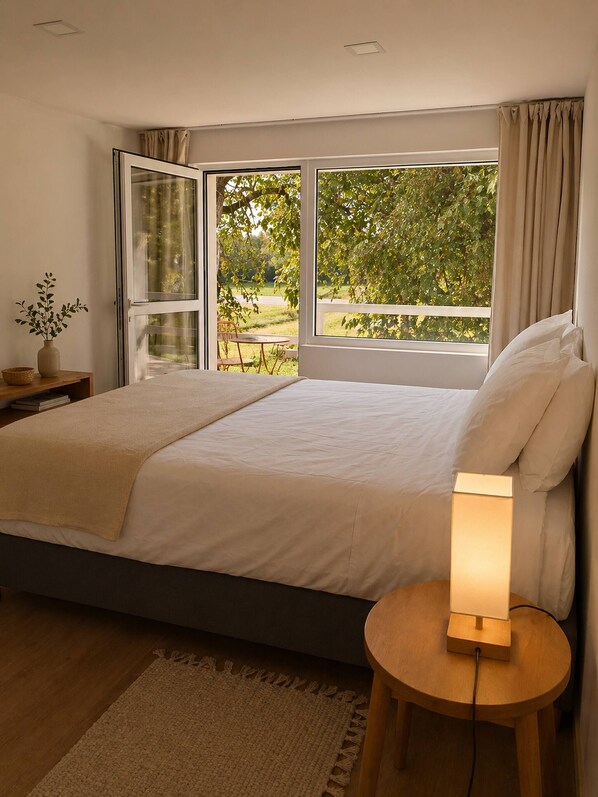 Un dormitorio con cama, una mesita de noche con lámpara, vistas al exterior con vegetación y una ventana con cortinas.
