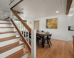 A staircase with wooden steps and white railings, a dining area with a table and chairs, and a large floral painting on the wall.