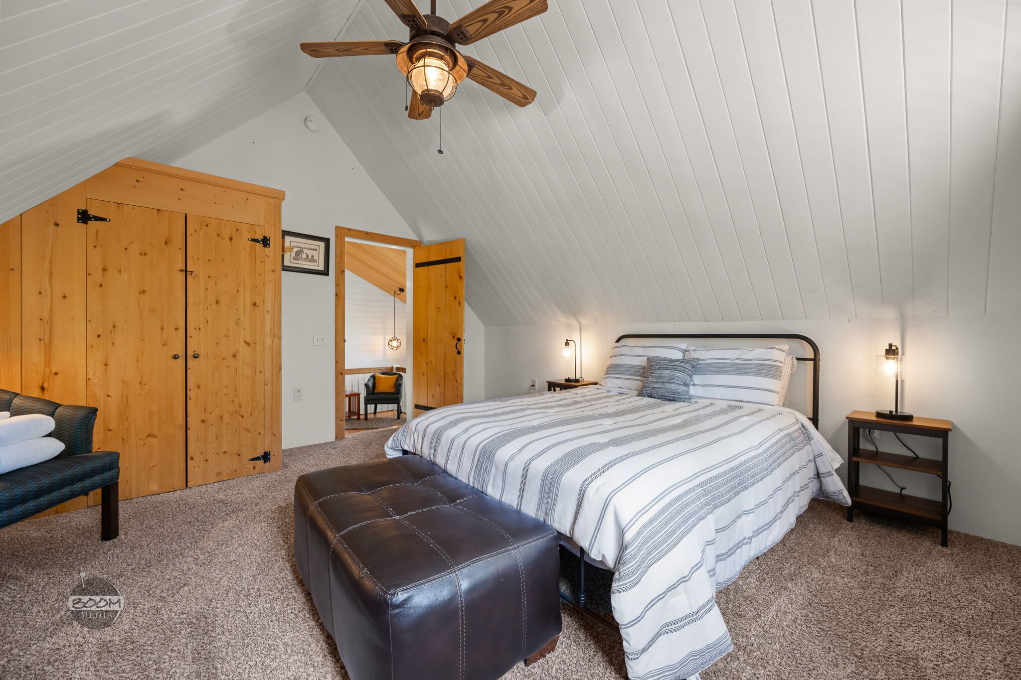 Ein Schlafzimmer mit geneigter Decke, einem Bett mit gestreifter Bettwäsche, einem Holzschrank, einer Bank und einem kleinen Tisch mit einer Lampe.