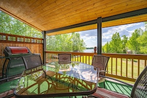 Un espace extérieur abrité, doté d’une table en verre, de chaises, d’un barbecue et offrant une vue sur un lac.