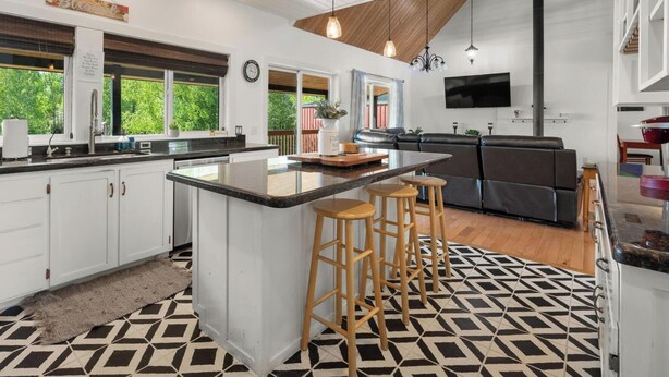 A modern kitchen with a central island, bar stools, a dining area with a sofa, and a large window with a view of greenery. - Lakeside Retreat- Charming 5 Bedroom, with Kayaks! (Wasilla)