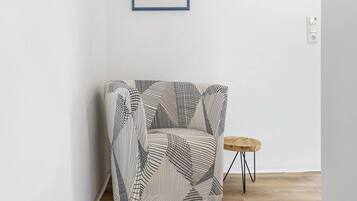 A corner room with a patterned armchair, a small wooden stool, and a framed picture on the wall.