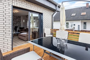 Une terrasse extérieure moderne, dotée d'une table en verre, de meubles en osier et offrant une vue sur la maison voisine.