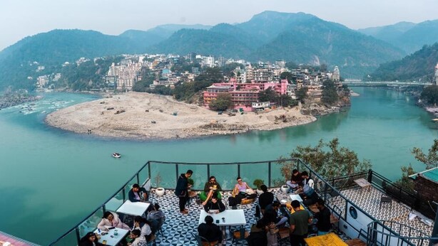 Een terras aan de rivier met tafels en stoelen, met uitzicht op een rivier en een stad met gebouwen en bergen op de achtergrond.