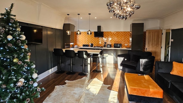 A modern living room with a large fur rug, a Christmas tree, and a dining area with a kitchen in the background. - Park View Penthouse Suite (Merseyside)
