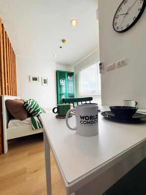 Een kamer met een bed, een witte tafel met een mok, een groen kastje en een wandklok.