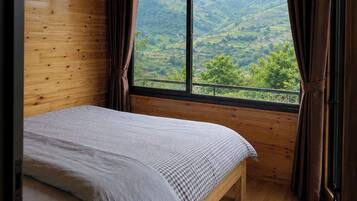 A bedroom with a wooden ceiling, a bed with a checkered bedspread, a window with curtains, and a view of green hills.