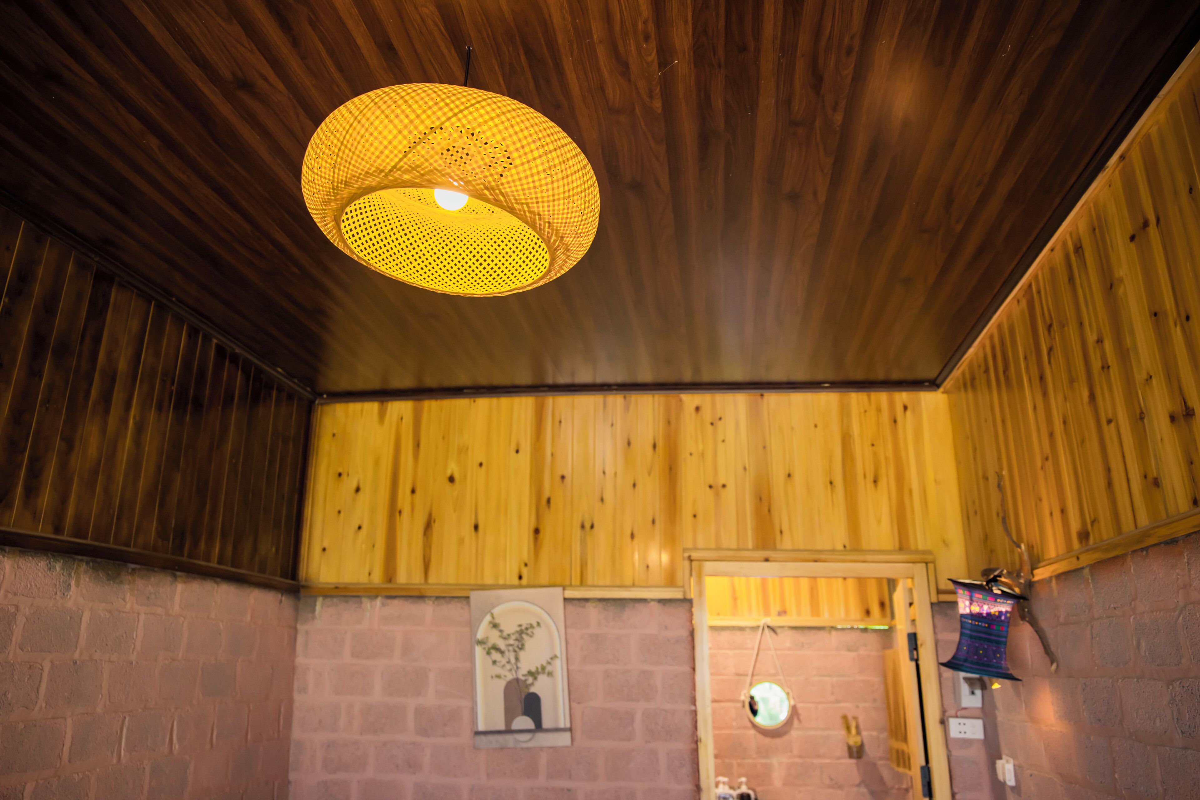 Quarto com paredes e teto de madeira, luminária pendente, porta e um pequeno espelho fixado na parede.