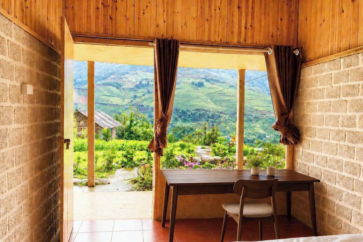 Una habitación con techo de madera, paredes de ladrillo, una mesa con sillas y vistas a un paisaje verde a través de ventanas grandes.