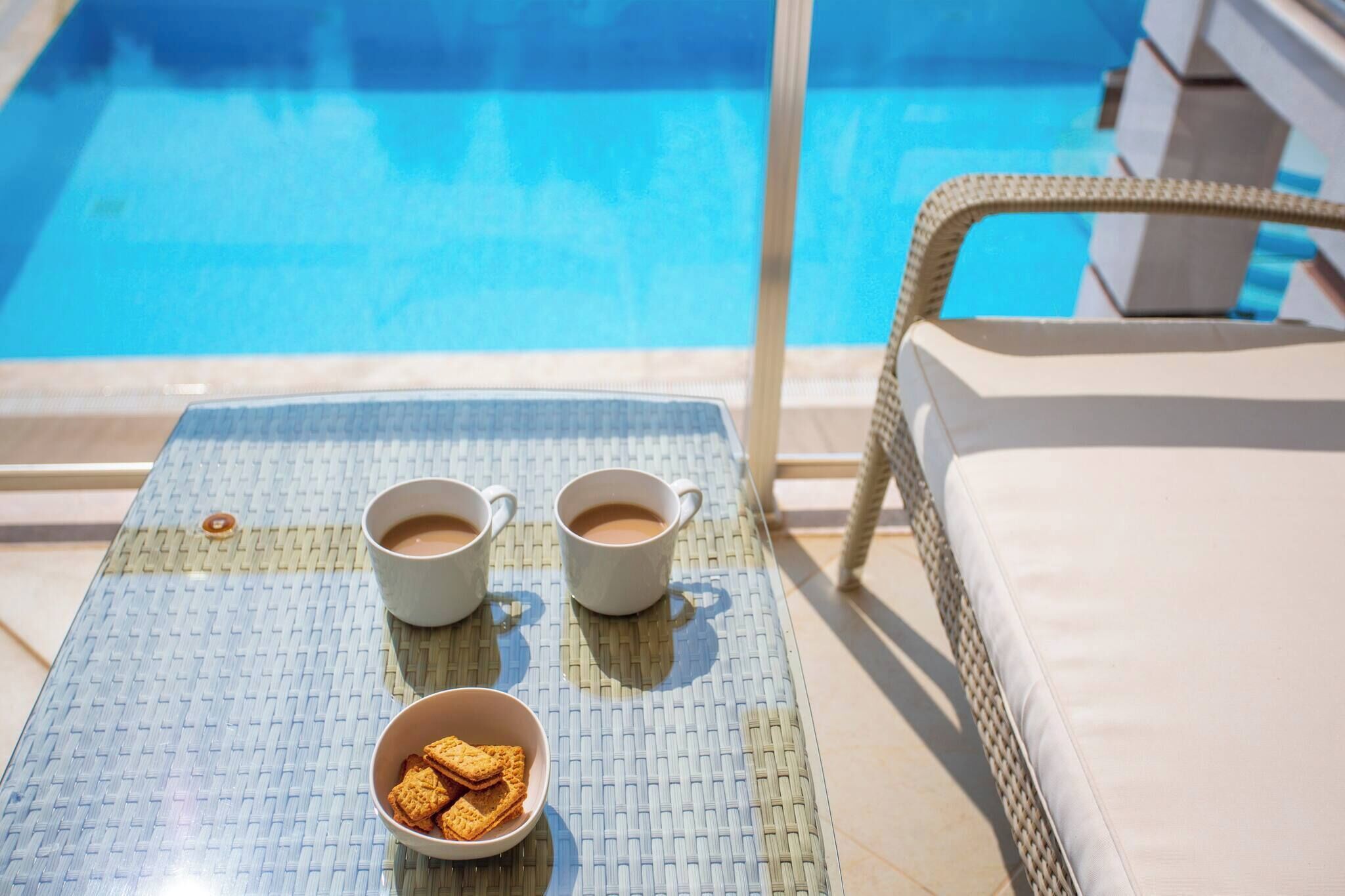 A poolside area with a table, two cups of coffee, and a bowl of cookies.