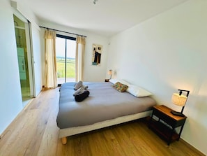 Quarto com cama, mesa de cabeceira, abajur e vista para o exterior.