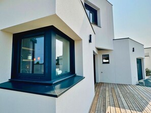 A modern building with a large window, a wooden deck, and a small outdoor area. - New architect-designed villa for 8 people, 4 bedrooms, enclosed garden, terrace, pétanque (Etaules, 17750)