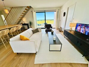 A modern living room with a sofa, a coffee table, a TV, and a staircase leading to a balcony with a view. - New architect-designed villa for 8 people, 4 bedrooms, enclosed garden, terrace, pétanque (Etaules, 17750)