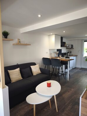 Un espace de vie moderne avec un canapé noir, deux petites tables rondes blanches, une cuisine en arrière-plan et un plancher en bois.