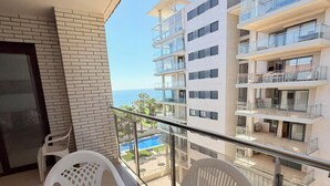 Un balcón con sillas blancas y una mesa, con vistas a una piscina y edificios cercanos.