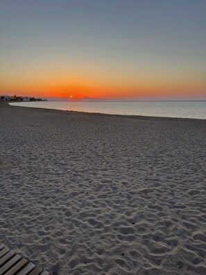 A beach with a sunset, sandy terrain, and distant buildings.