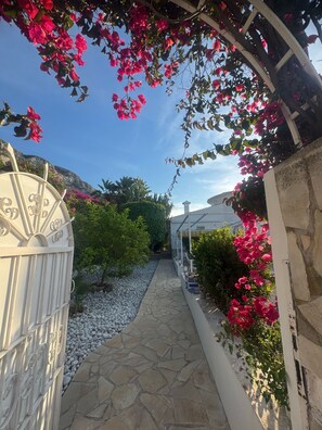 Un camino de piedra que lleva a una puerta blanca con un diseño decorativo, rodeado de follaje y flores rosadas.
