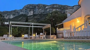 Una piscina con una pérgola, sillas de descanso y montañas al fondo.