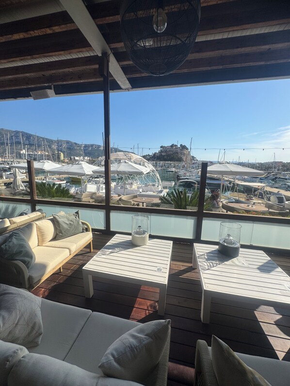 Una terraza con mesas al aire libre, vista a un puerto deportivo y una montaña al fondo.