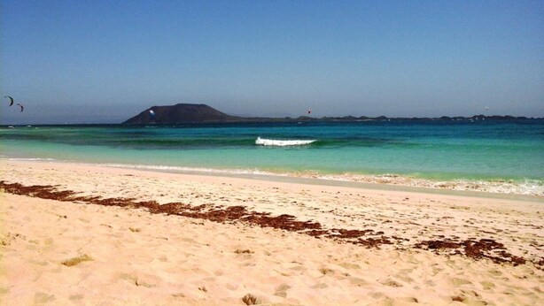 A beach with clear turquoise water, a sandy shore, and a distant island. - Lovely   Apt  Siesta (Corralejo)