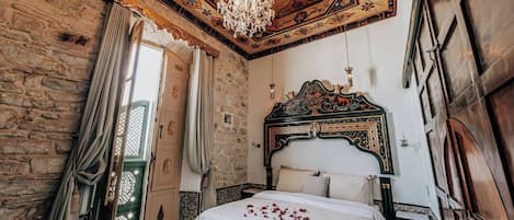Una habitación con pared de piedra, cama de madera con cabecera decorativa, candelabro y puerta verde.