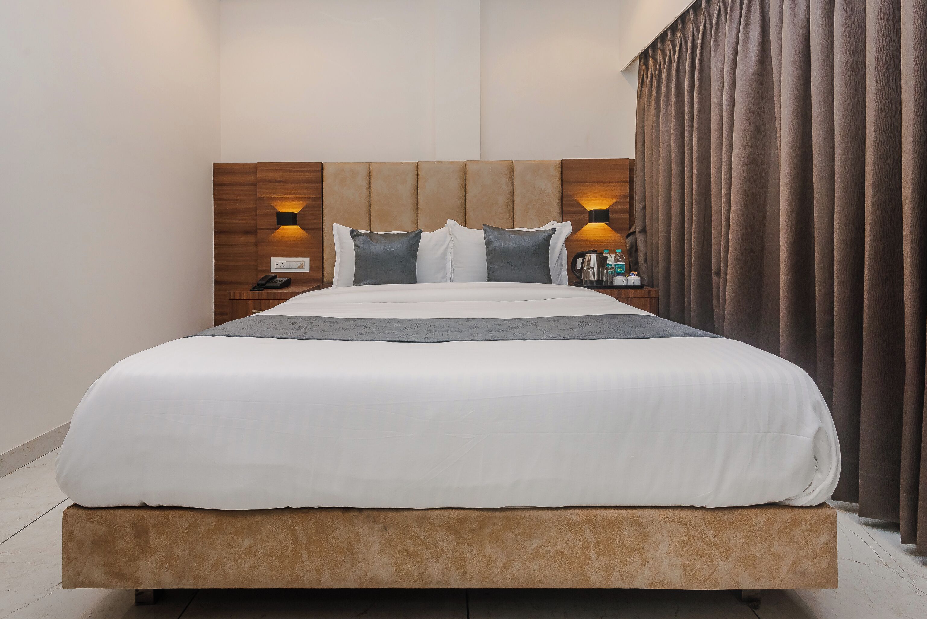 A modern hotel room with a large bed, wooden headboard, two bedside lamps, and a nightstand with toiletries.