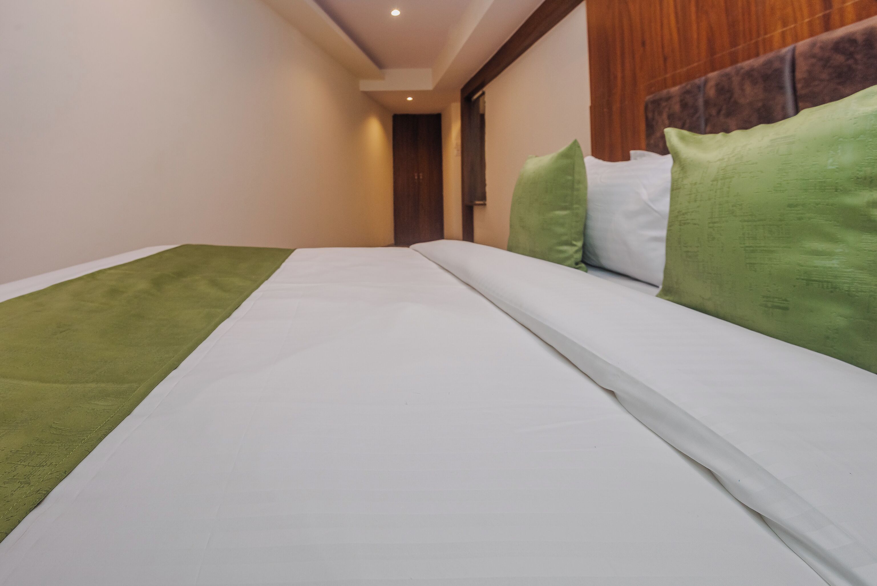 Habitación de hotel con cama, almohadas verdes y blancas, cabecera de madera y pared blanca.