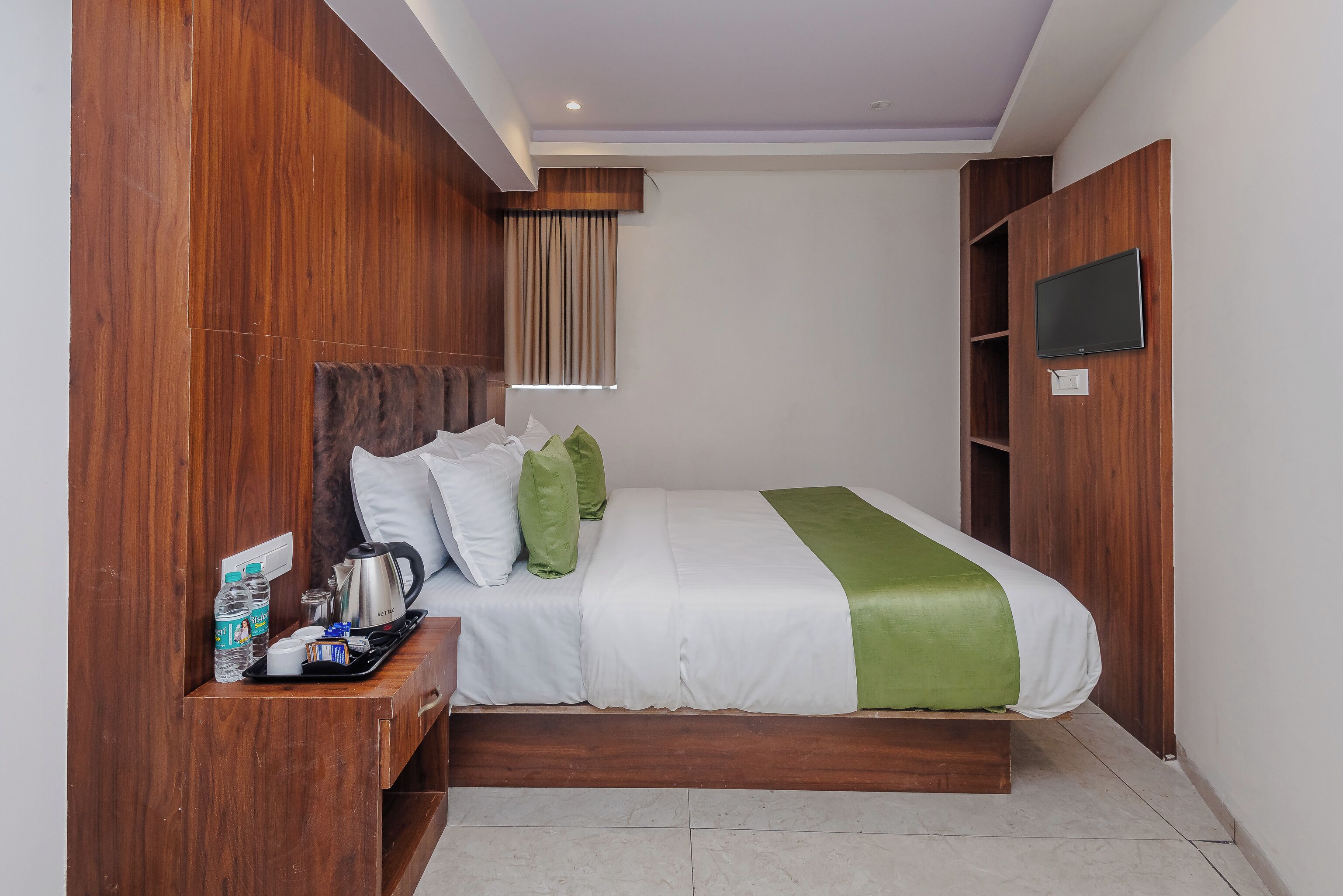 Habitación de hotel ordenada con una cama, cabecero de madera, una mesita con un hervidor y un televisor montado en la pared.